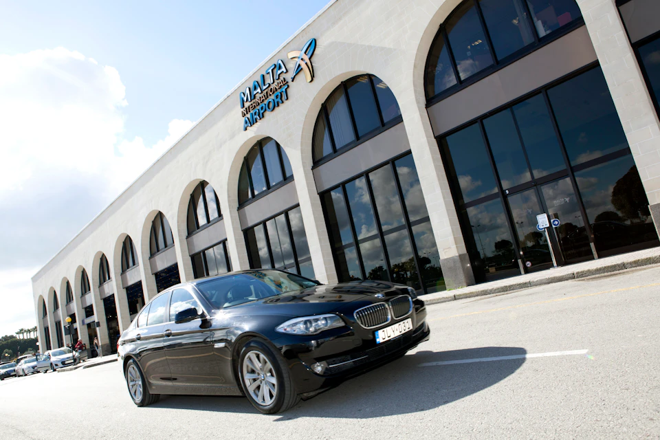A black car is parked in front of Malta International Airport's building, featuring large arched windows and reflective glass.