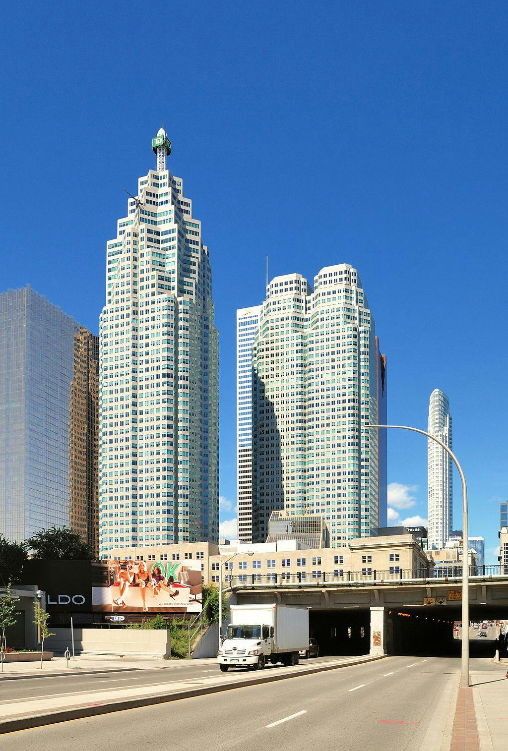 Grattacieli alti sotto un cielo azzurro, un cartellone pubblicitario accanto a una strada e un camion vicino a un sottopassaggio.