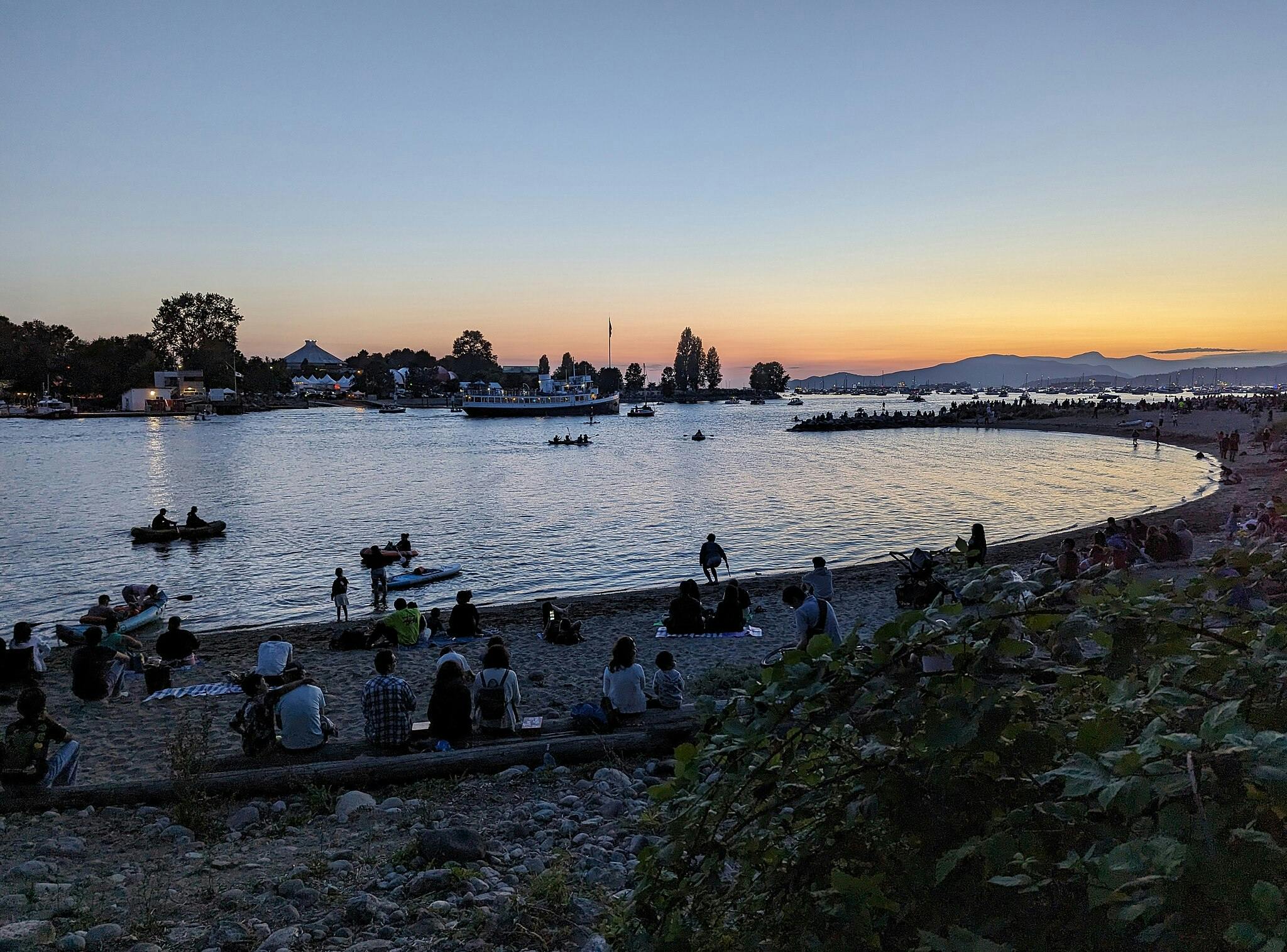 Menschen an einem Strand in der Abenddämmerung, mit Booten auf ruhigem Wasser und einem Sonnenuntergang im Hintergrund.