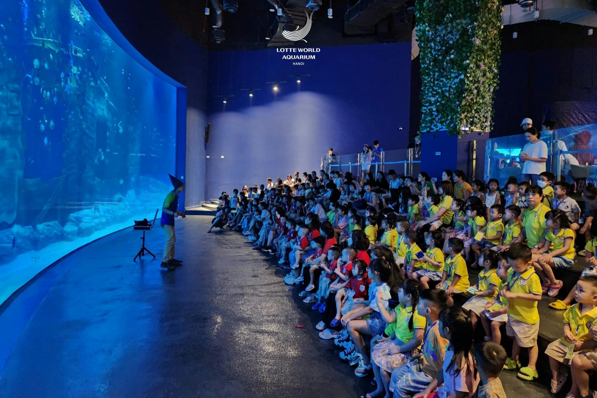 一大群穿着黄色T恤的儿童在乐天世界水族馆观看演讲，一名工作人员在前面发言。