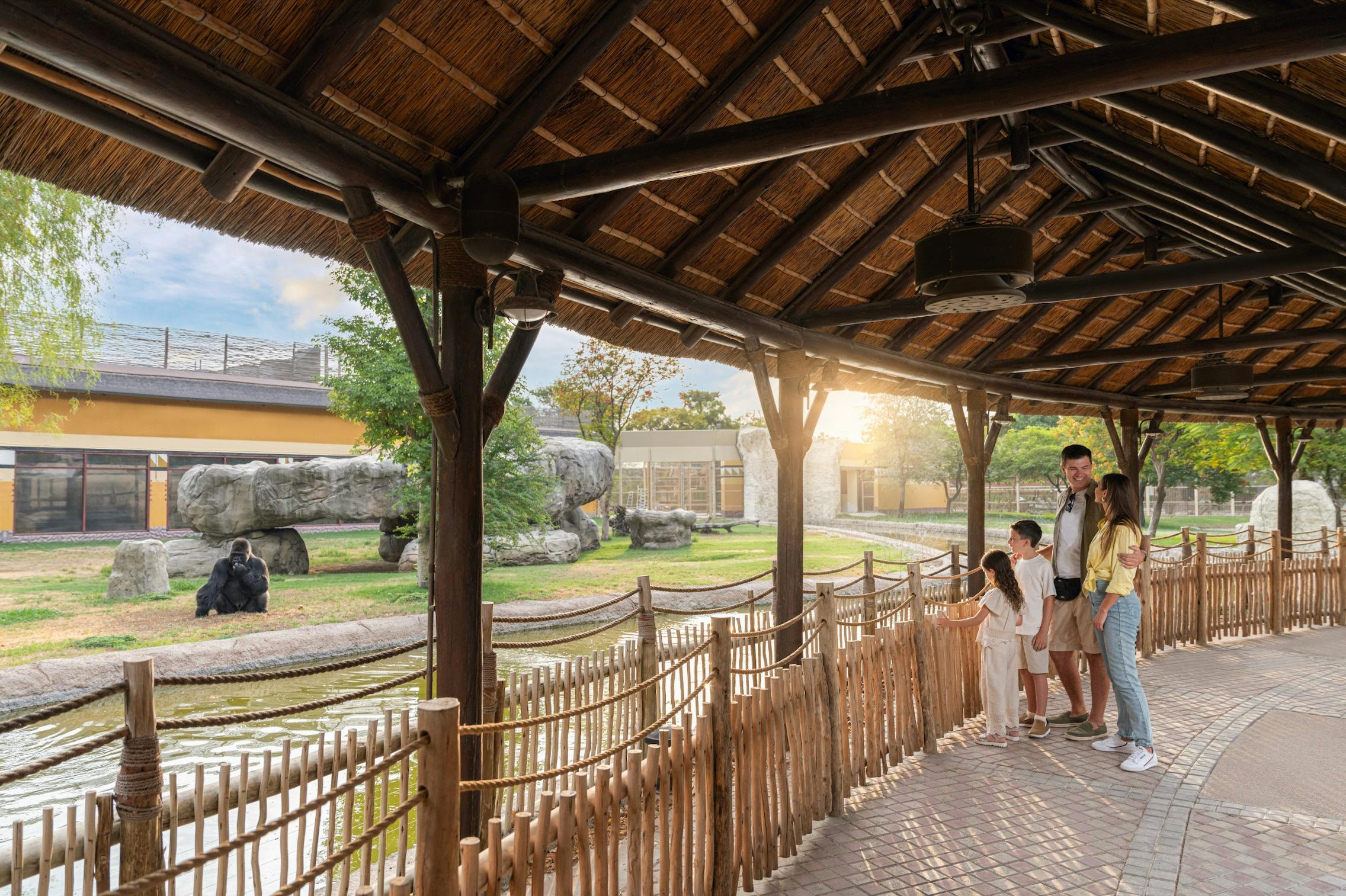 Μια τετραμελής οικογένεια στέκεται κάτω από μια αχυρένια δομή στέγης, κοιτάζοντας έναν υπαίθριο περίβολο ζωολογικού κήπου με δέντρα και βράχια στο παρασκήνιο.