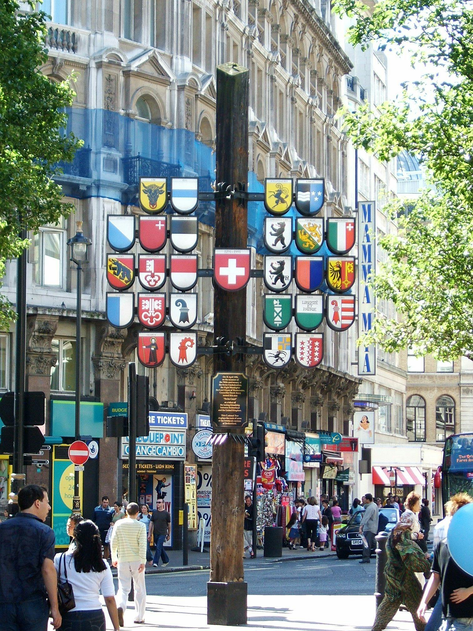 Street scene with people walking, decorative shields on a pole, shops, and a theater sign reading "MAMMA MIA!" in the background.