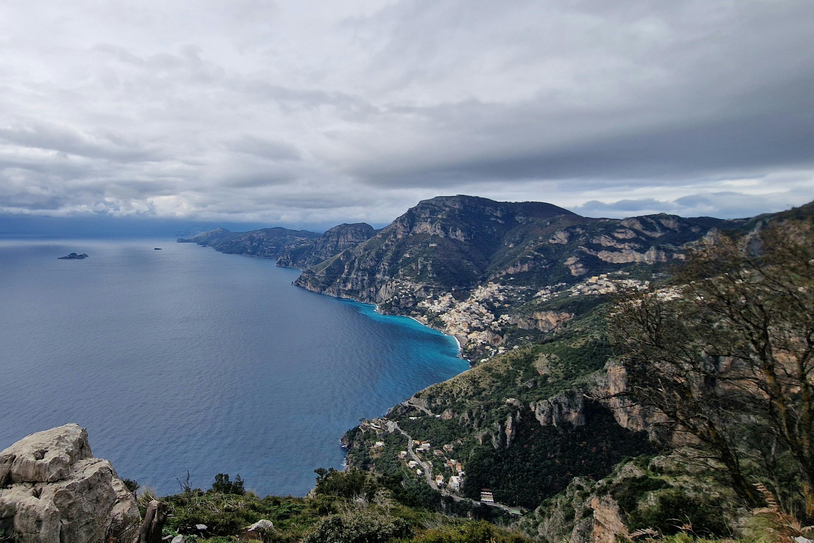 A coastal mountain landscape with a village nestled along the shoreline, surrounded by blue sea and under a cloudy sky.