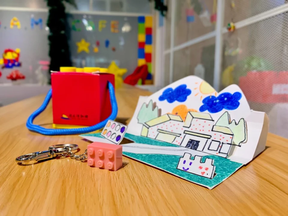 A colorful drawing of a house on a table with a keychain, a red box, and a brick-shaped charm in the foreground.