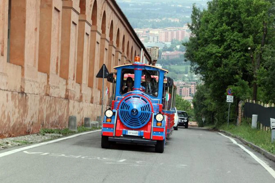A small blue and red tourist train driving on a road beside an old beige building with arches, with trees and city in the background.
