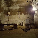 Dimly lit underground chapel with stone benches, multiple crosses, candles, religious icons, and a fenced-off area on the right.