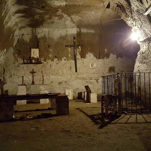 Dimly lit underground chapel with stone benches, multiple crosses, candles, religious icons, and a fenced-off area on the right.