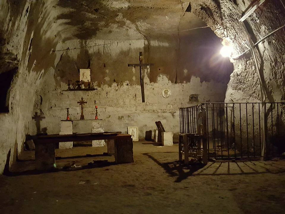 A dimly lit underground chapel with stone benches, several crosses, candles, religious icons, and a fenced-off area on the right.
