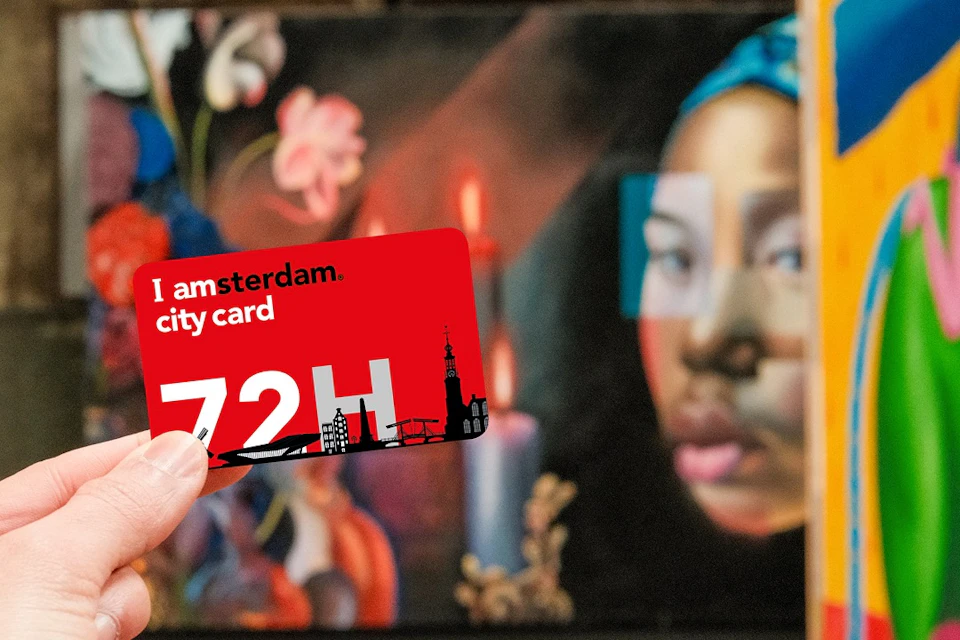 A hand holding an Amsterdam city card with a red background in front of a painting featuring a woman and flowers.