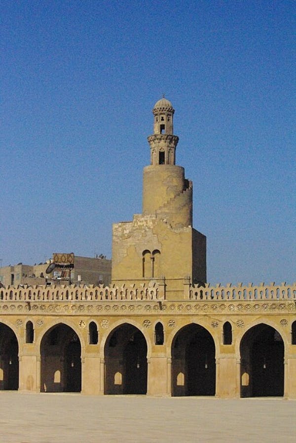 A historic mosque with an arched colonnade and a spiral minaret against a clear blue sky.