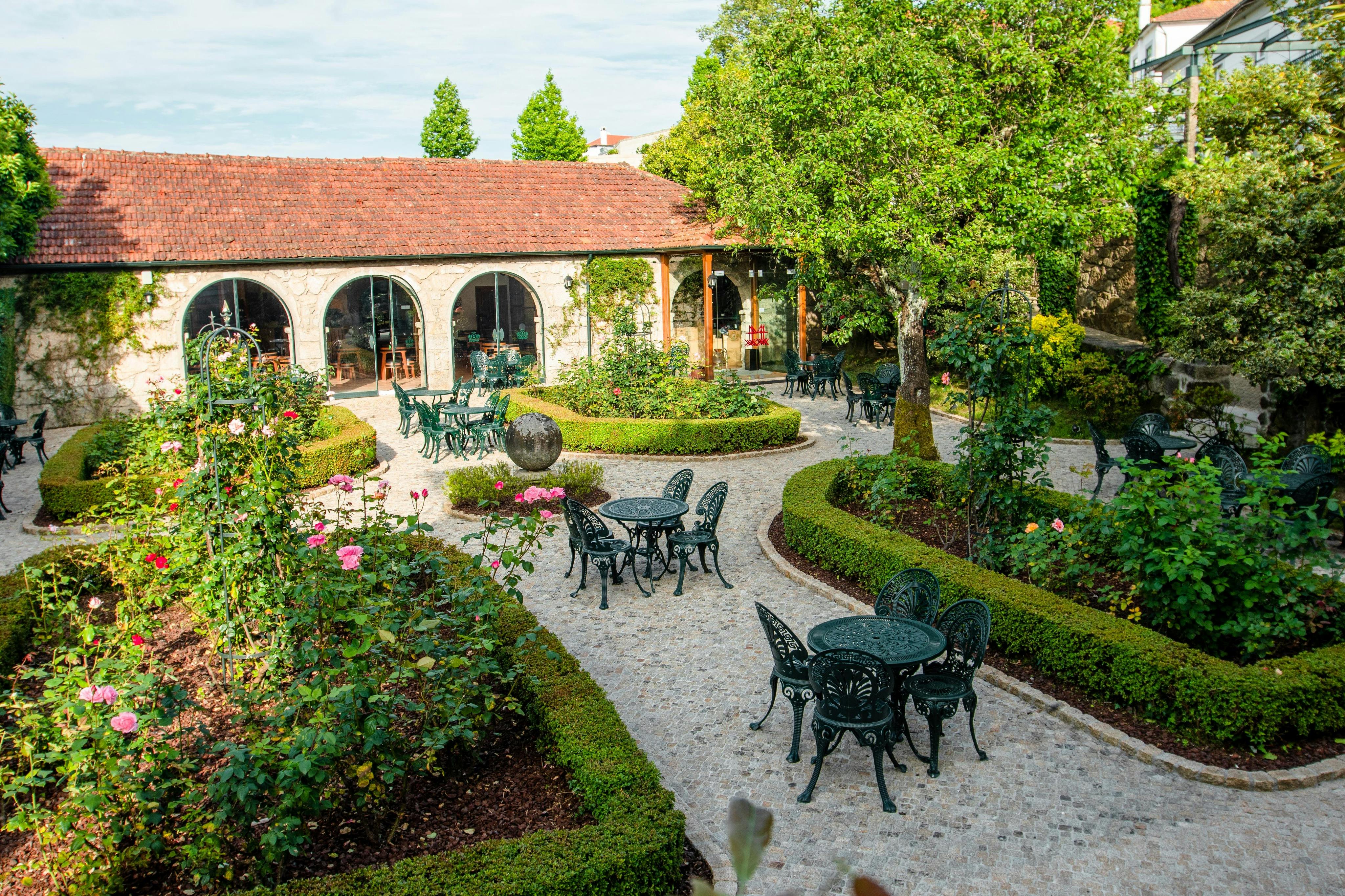 Nádvoří s četnými zelenými kovovými stoly a židlemi, úhledně upravenými živými ploty, rozkvetlými růžovými keři a budovou s klenutými okny.