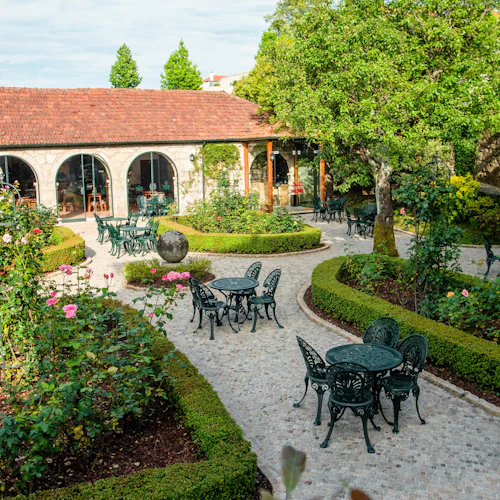 En innergård med många gröna metallbord och stolar, prydligt trimmade häckar, blommande rosenbuskar och en byggnad med välvda fönster.