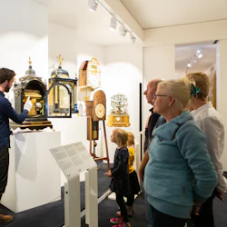 En guide i en blå skjorta visar antika klockor för en grupp vuxna och barn på ett museum.