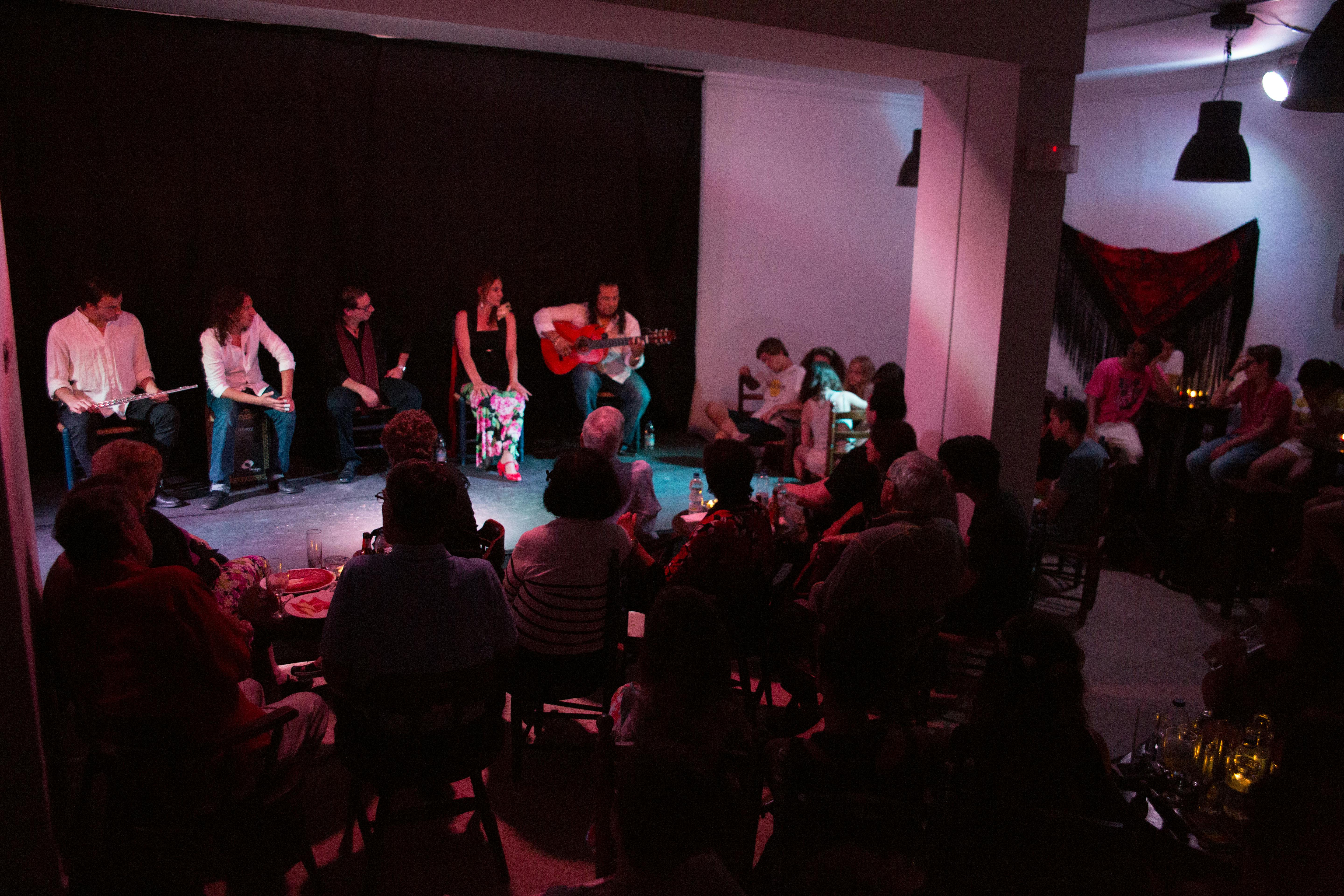 A man plays guitar on a stage with a small audience watching in a dimly lit room. Other performers sit on stage.