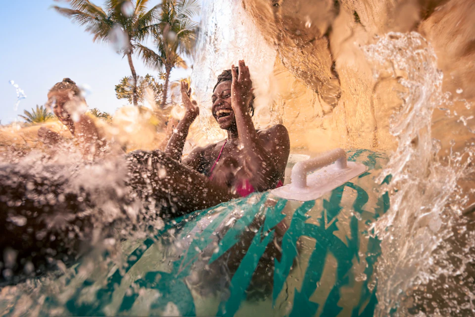 A person enjoying a water slide, splashing through water on an inflatable tube, with palm trees in the background.