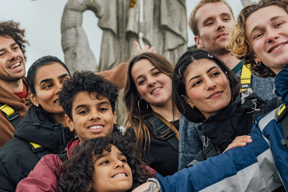 A group of people in safety harnesses take a selfie near a stone statue with wings, outdoors with a city view in the background.