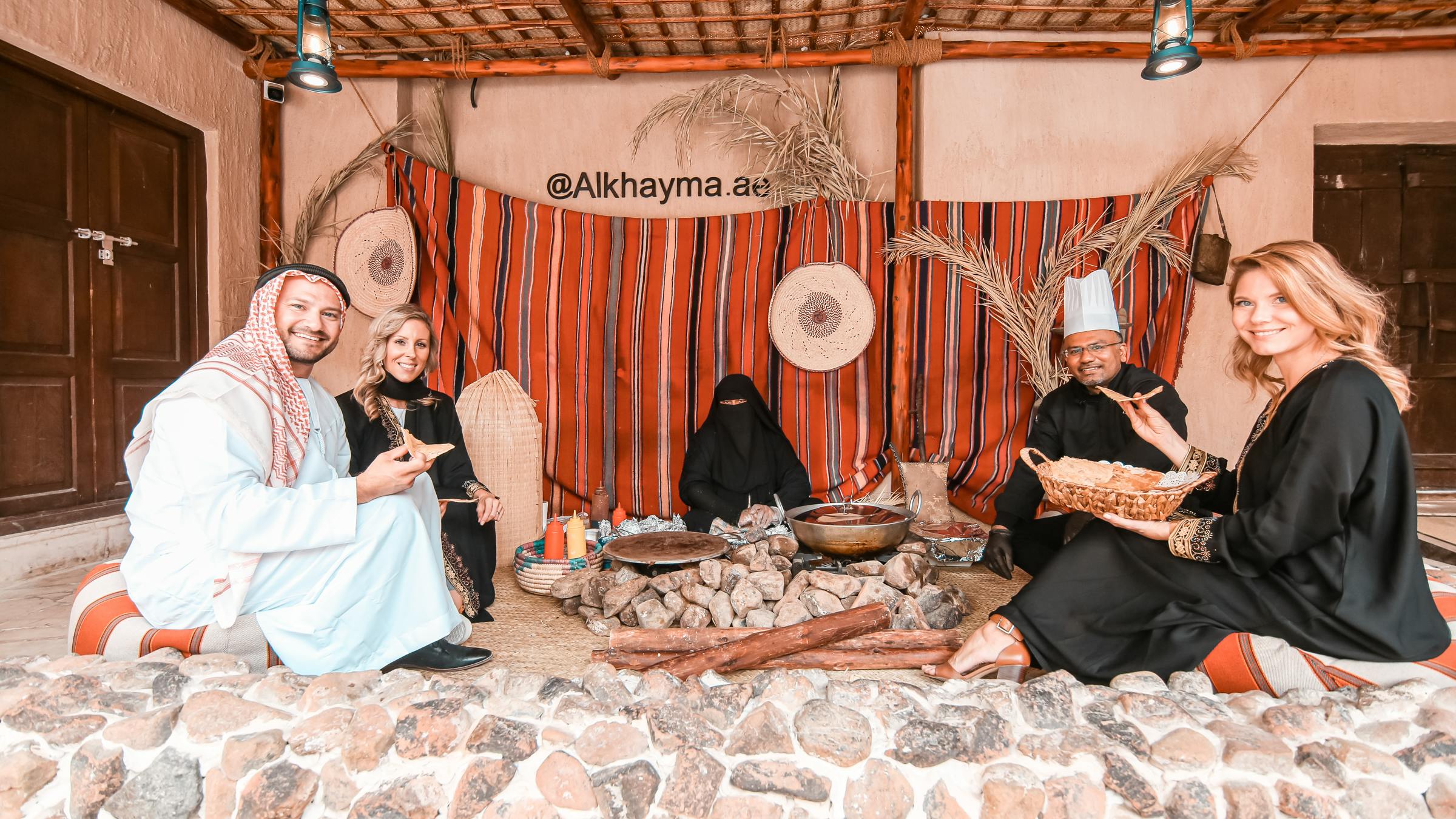 Five people in traditional clothing sitting around a stone fire pit with food, under a wooden shelter with striped fabric backdrop.