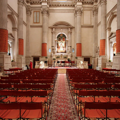 Utsmyckad inredning med rader av röda stolar, stora pelare, religiösa statyer och altare med målning och blommor. Ensamstående person i röd mantel.