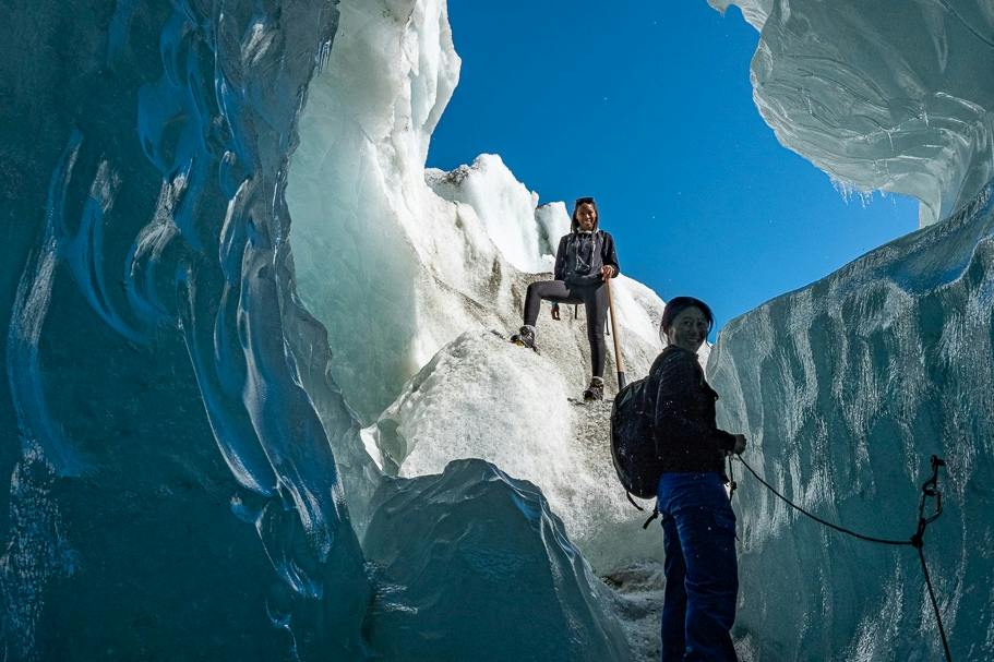 Two people climbing through a glacier crevasse, surrounded by ice walls under a clear blue sky.