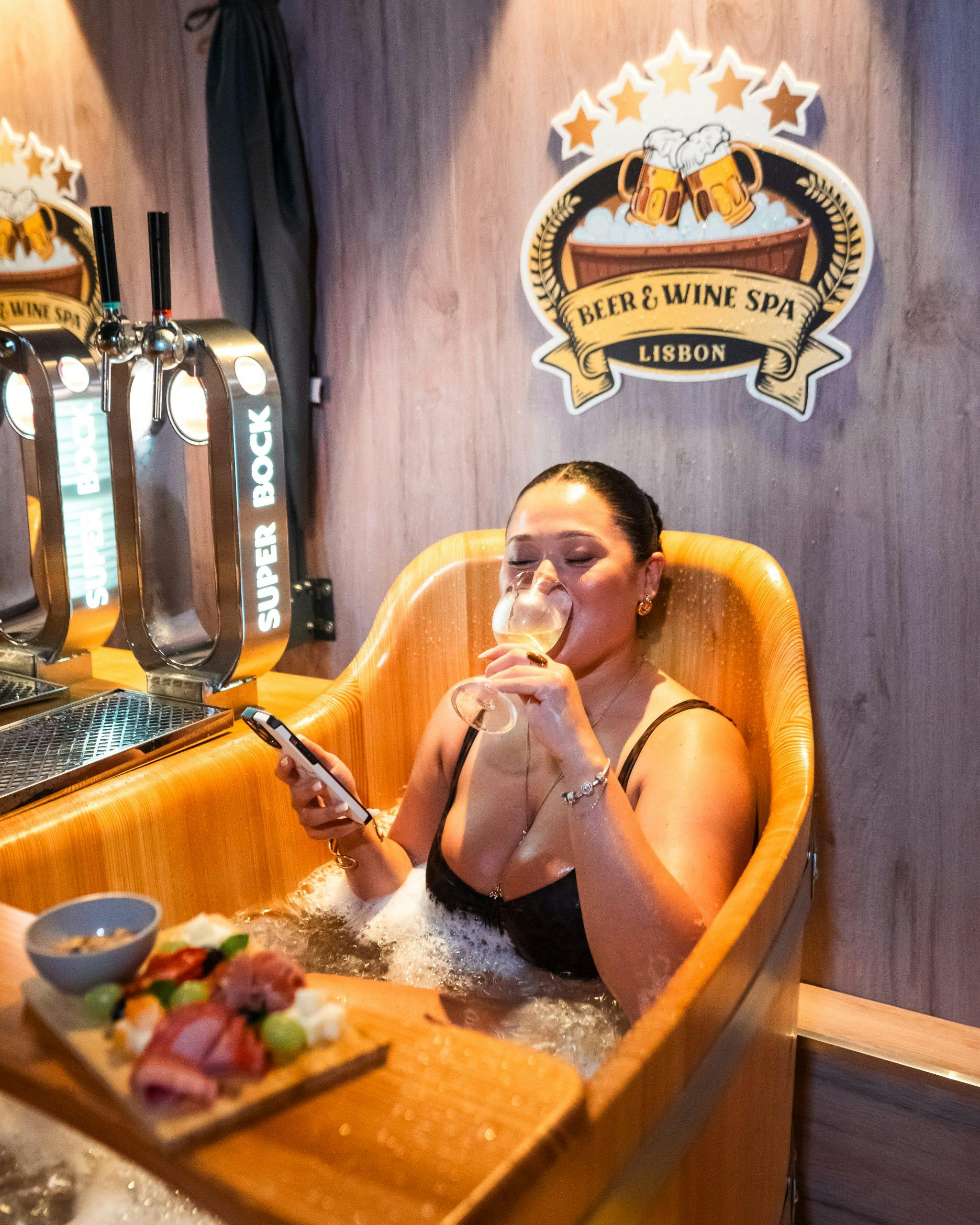 Μια γυναίκα χαλαρώνει σε μια ξύλινη μπανιέρα σε ένα σπα μπύρας και κρασιού, πίνοντας κρασί και κρατώντας ένα τηλέφωνο, με φαγητό και βρύσες μπύρας κοντά.
