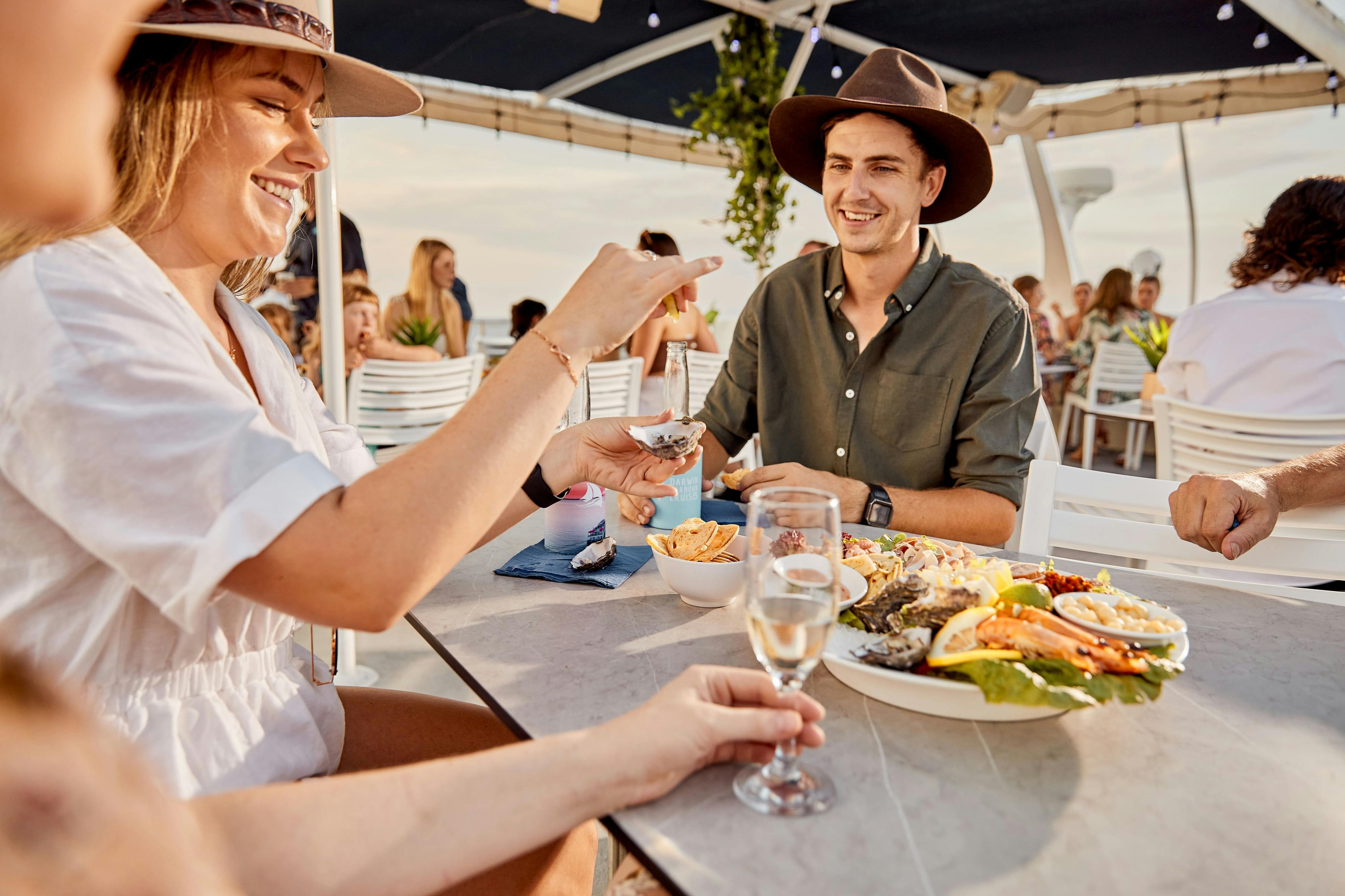 茶色の帽子をかぶり、緑色のシャツを着た男性が、シーフードとドリンクを並べた屋外のテーブルに座って、人々に囲まれながら微笑んでいます。