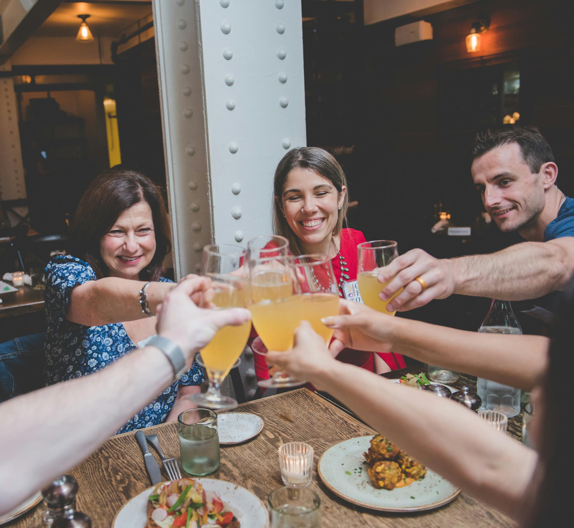 Smiling friends toasting with drinks around a table with plates of food and a candle in a warmly lit restaurant.