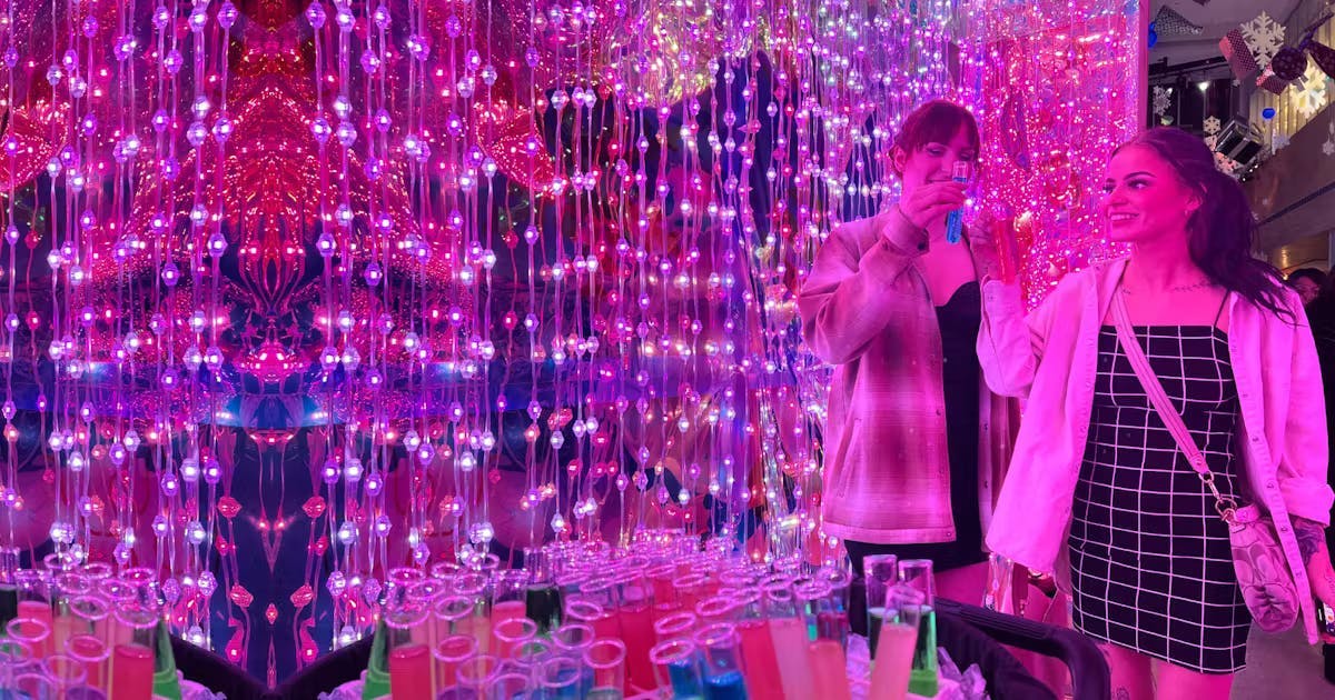 다채롭고 빛나는 줄 조명으로 둘러싸인 테스트 튜브에서 음료를 즐기는 두 여성이 서로에게 미소를 짓고 있습니다.