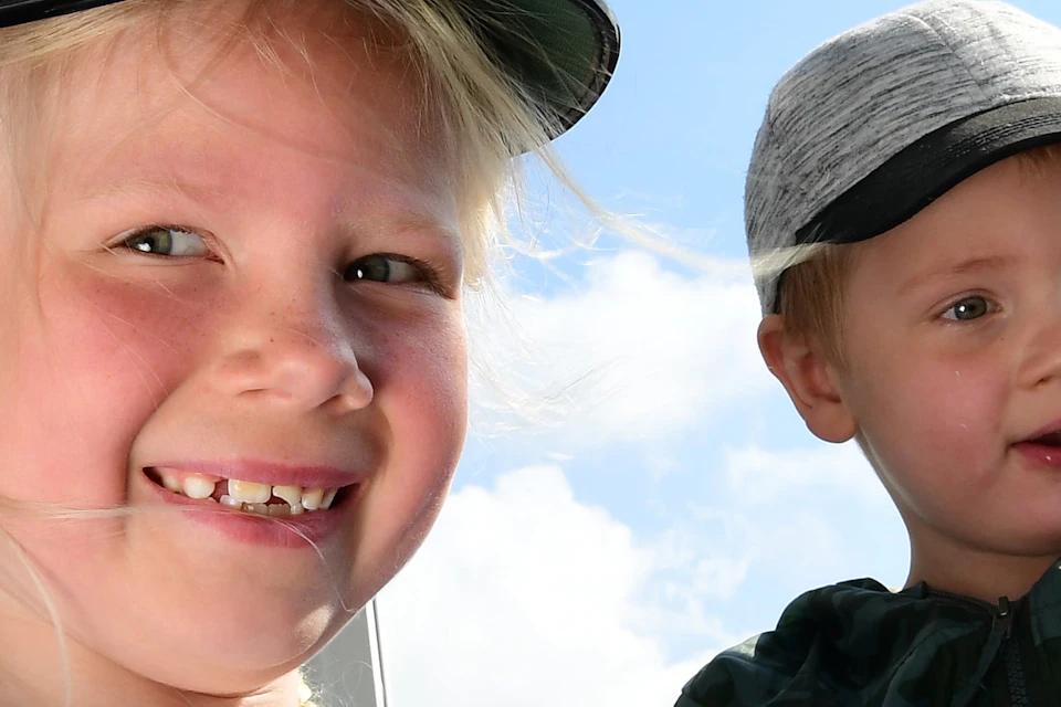 Two children in a bright setting; one wears a sailor's hat and blue shirt, smiling, the other in a cap and camo jacket.