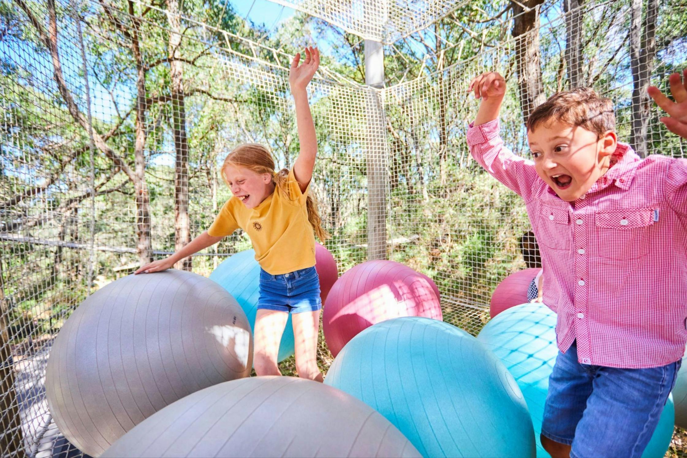 В солнечный день ребенок играет на разноцветных шариках для упражнений в игровой зоне, окруженной деревьями.
