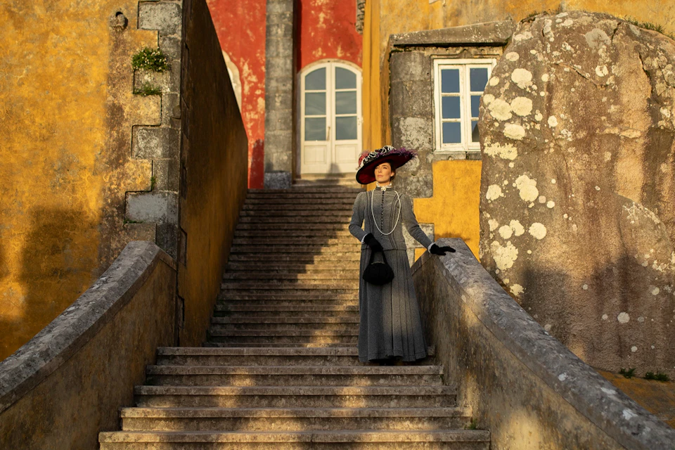 A person in vintage attire stands on stone stairs outside a colorful building with a red door and arched windows.