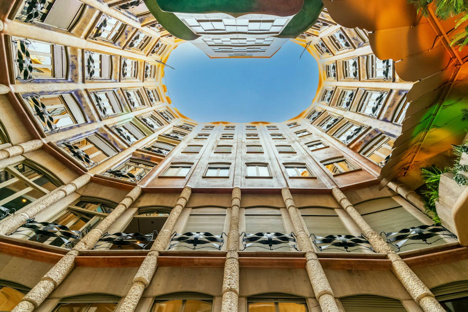 Upward view of a circular building courtyard with windows and balconies, opening to a blue sky.