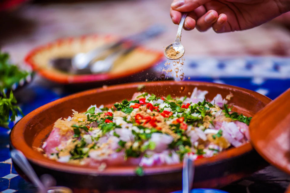 A hand sprinkling spices over a dish with chopped vegetables and herbs in a brown bowl. Blurred utensils in the background.