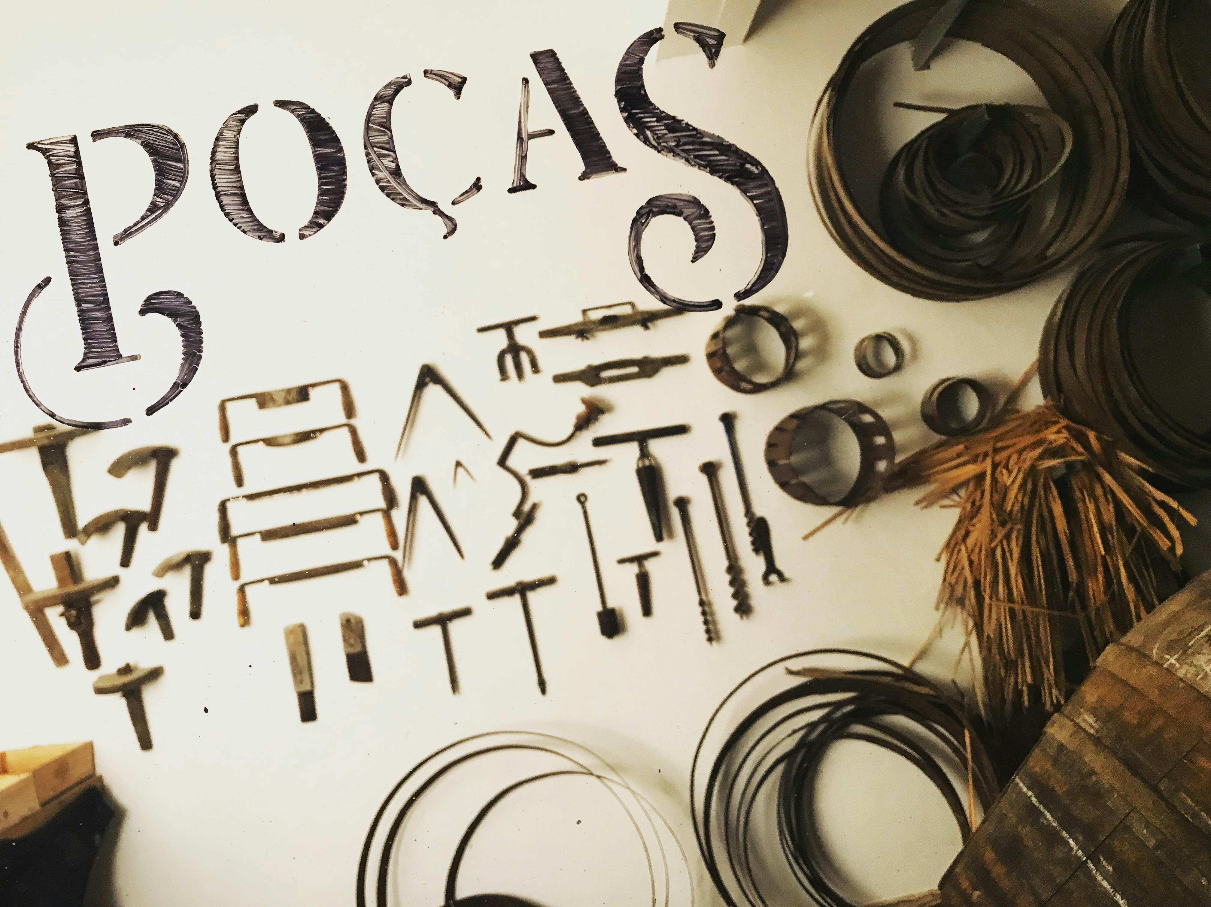 白い表面に配置された様々な金属工具と円形の物体、その上に部分的に見えるテキスト「POÇAS」。