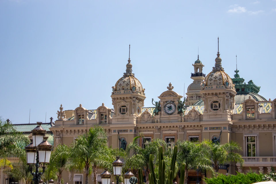 A historic building with ornate architecture, domes, a clock, and palm trees in the foreground under a clear sky.