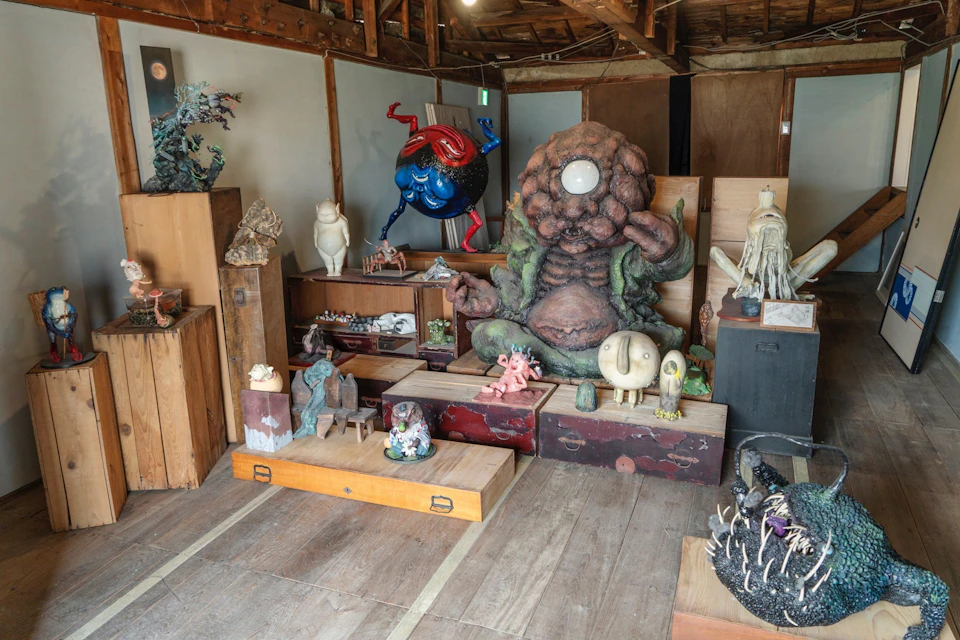 A room filled with various artistic sculptures, including a large seated creature, displayed on wooden boxes and tables.