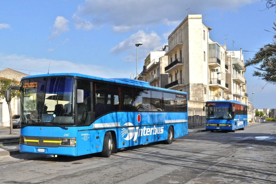 Two blue Interbus buses on a street beside a multi-story building under a partly cloudy sky.