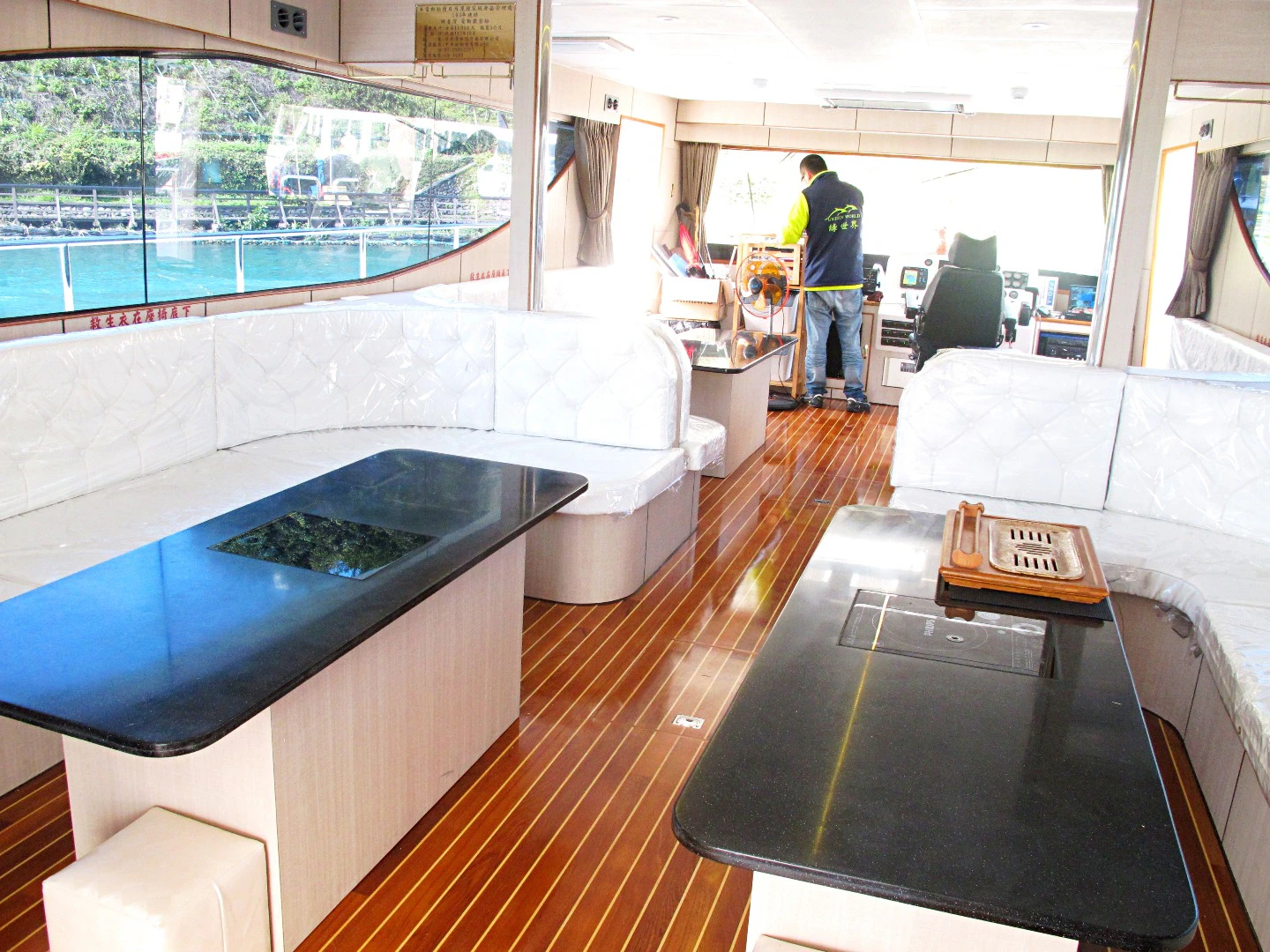 Interior of a modern boat with wooden flooring, white seating, black countertops, and a person working at the helm.