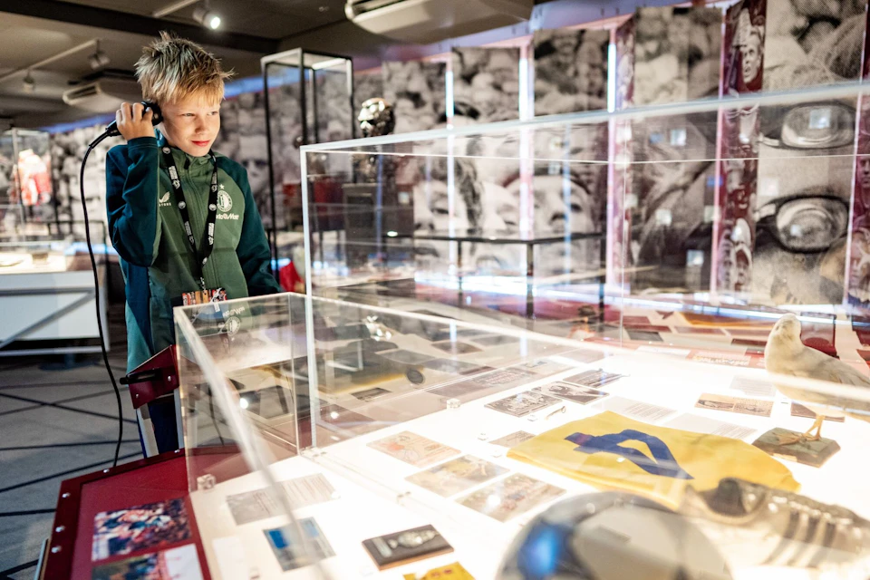 A young boy is interacting with an exhibit in a museum, holding an audio guide, near a glass display case containing various items.