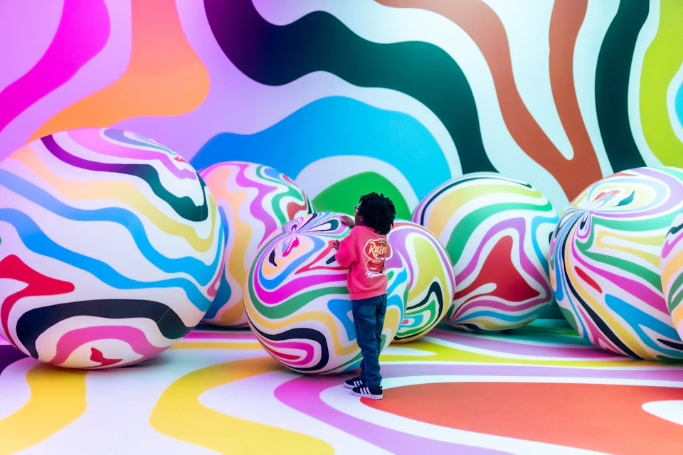 A child in a pink jacket interacts with large colorful striped balls in a vibrant room with wavy patterns on the walls and floor.