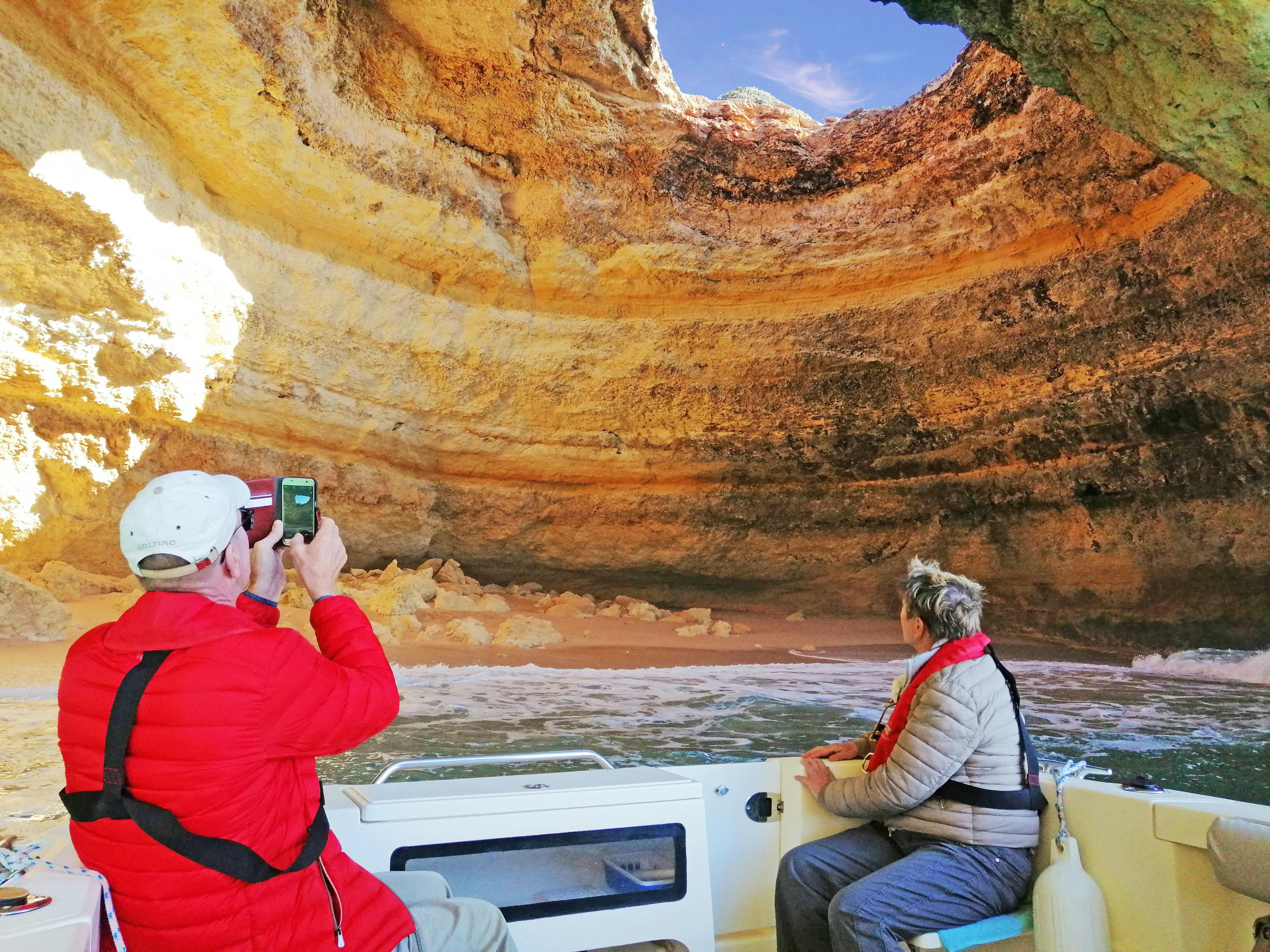 Two people in jackets on a boat inside a cave with layered rock walls. One is taking a photo with a phone, the other is looking around.