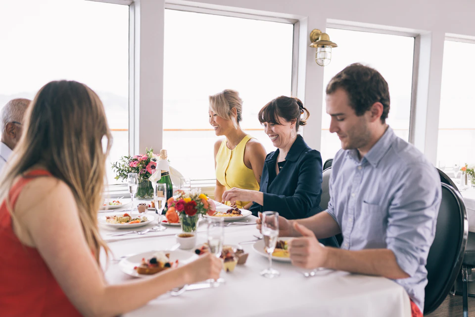 Four people dining together at a table with flowers and plates of food on a boat or by large windows.