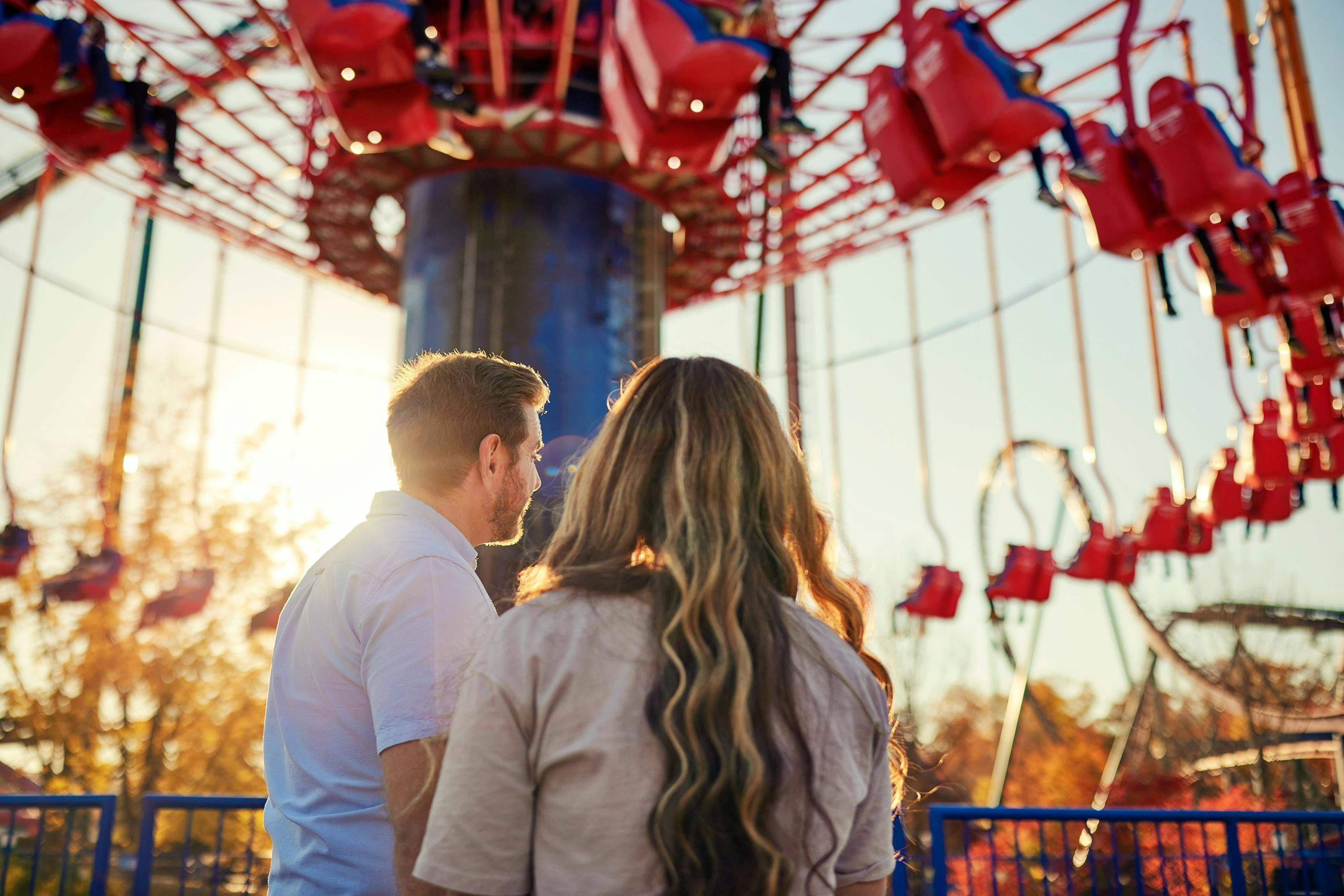 Два человека, стоящие перед красным парком развлечений, едут с распашными стульями, с солнечным светом и падающими деревьями на заднем плане.