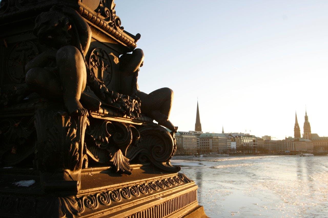 Scultura ornata con figure umane su un ponte che si affaccia su un fiume, con un paesaggio urbano e un campanile sullo sfondo.