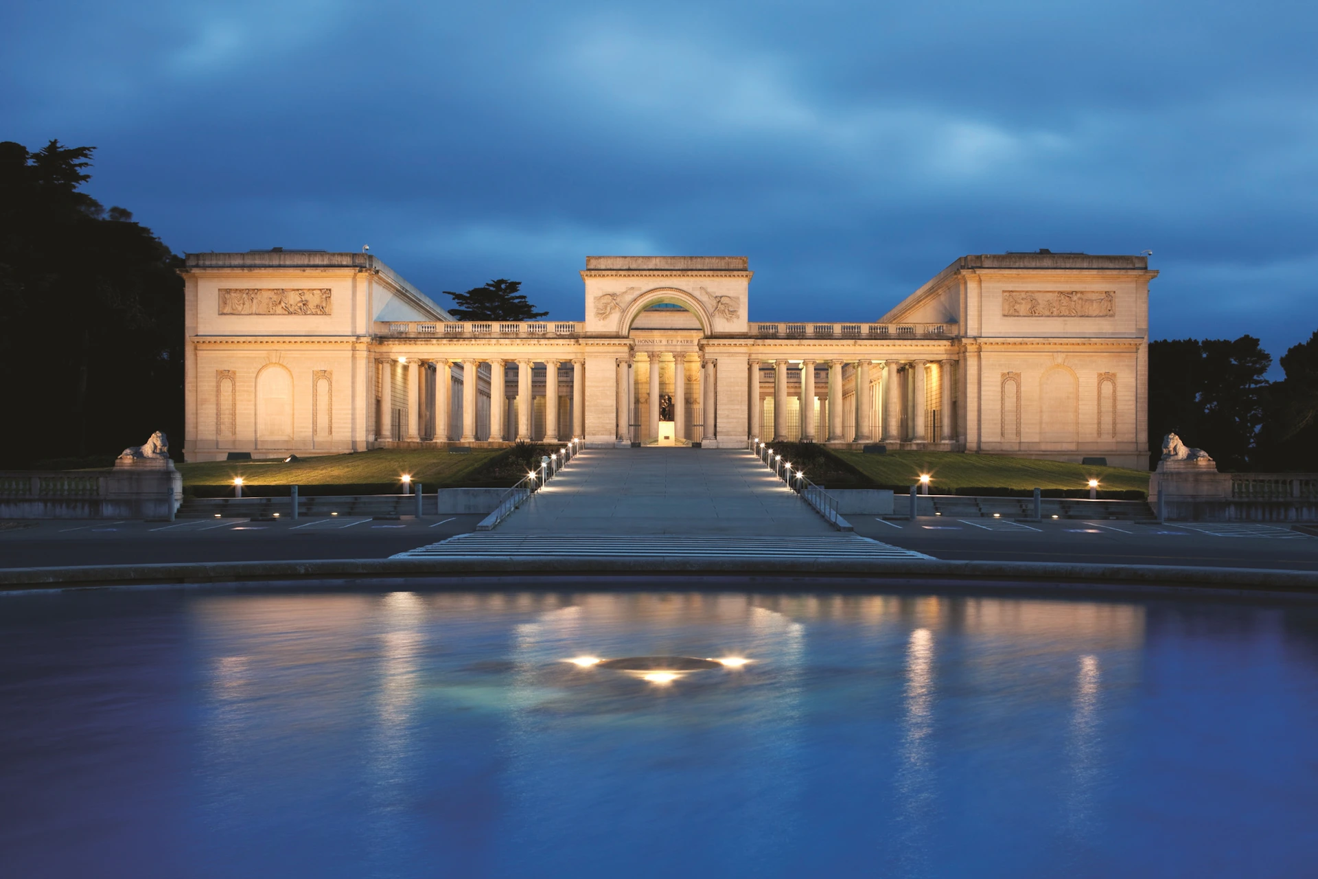 Illuminated classical building with columns and detailed archway, viewed from across a calm reflecting pool during twilight.