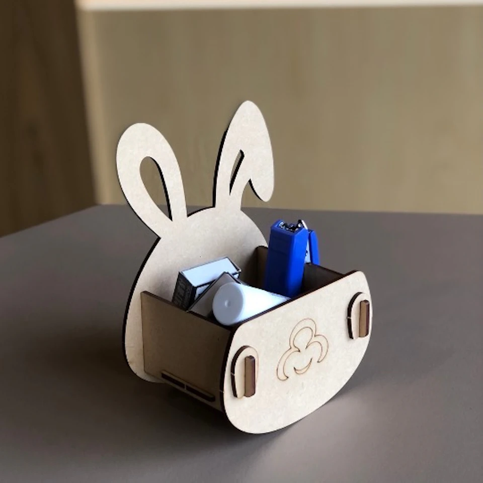 A wooden bunny-shaped organizer holding small items such as a blue stapler and other stationery on a table.