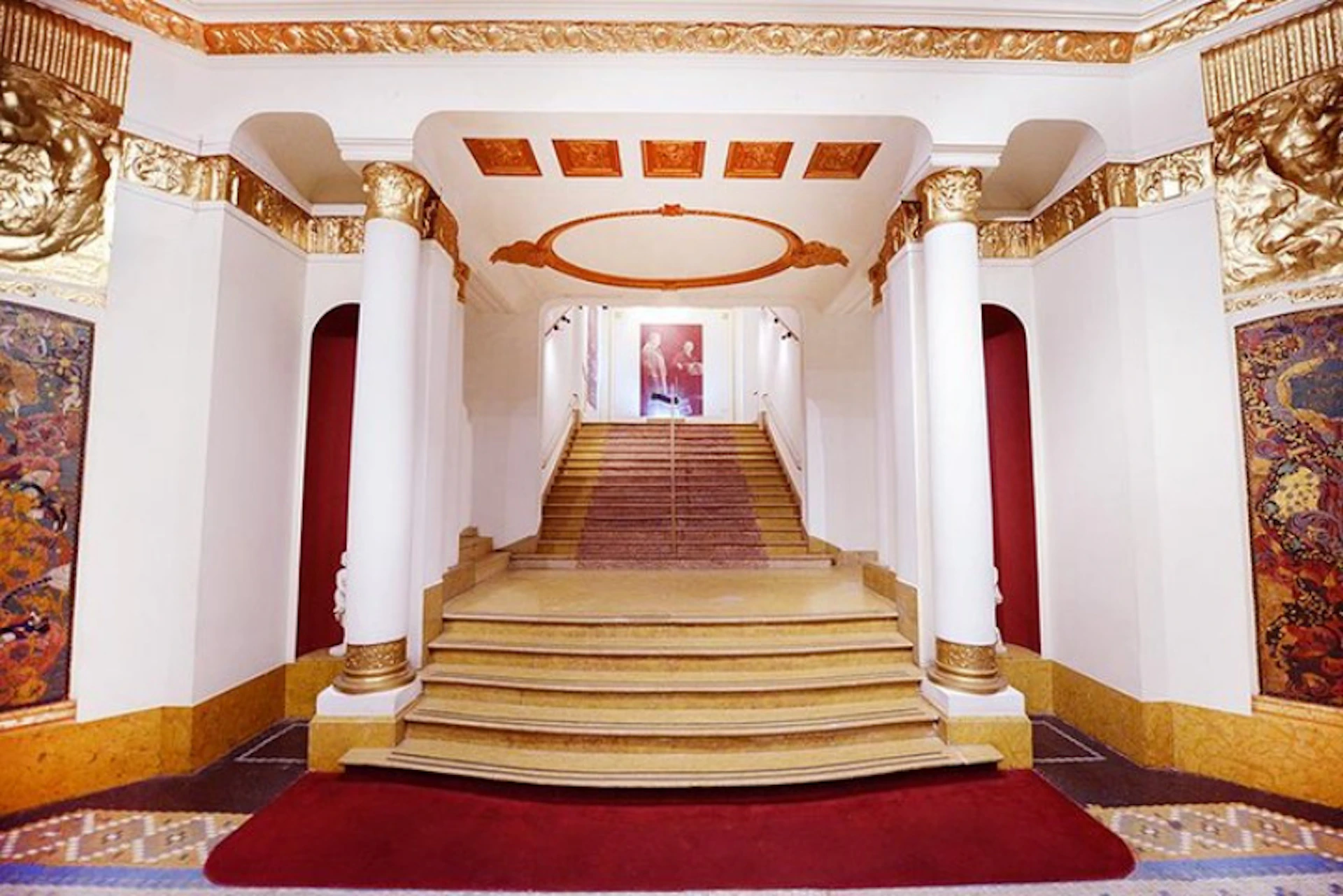 Elegant staircase with white and gold columns, red carpet, and a decorative ceiling leading to a portrait at the top.