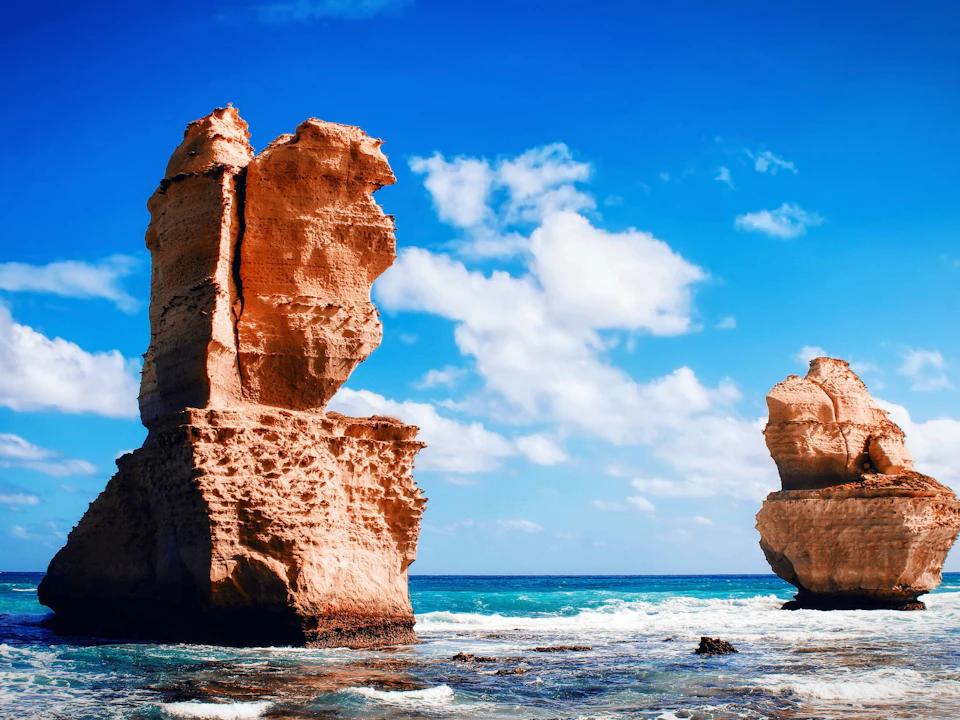 Two tall, weathered rock formations stand in the ocean with waves crashing against them under a bright blue sky with scattered clouds.