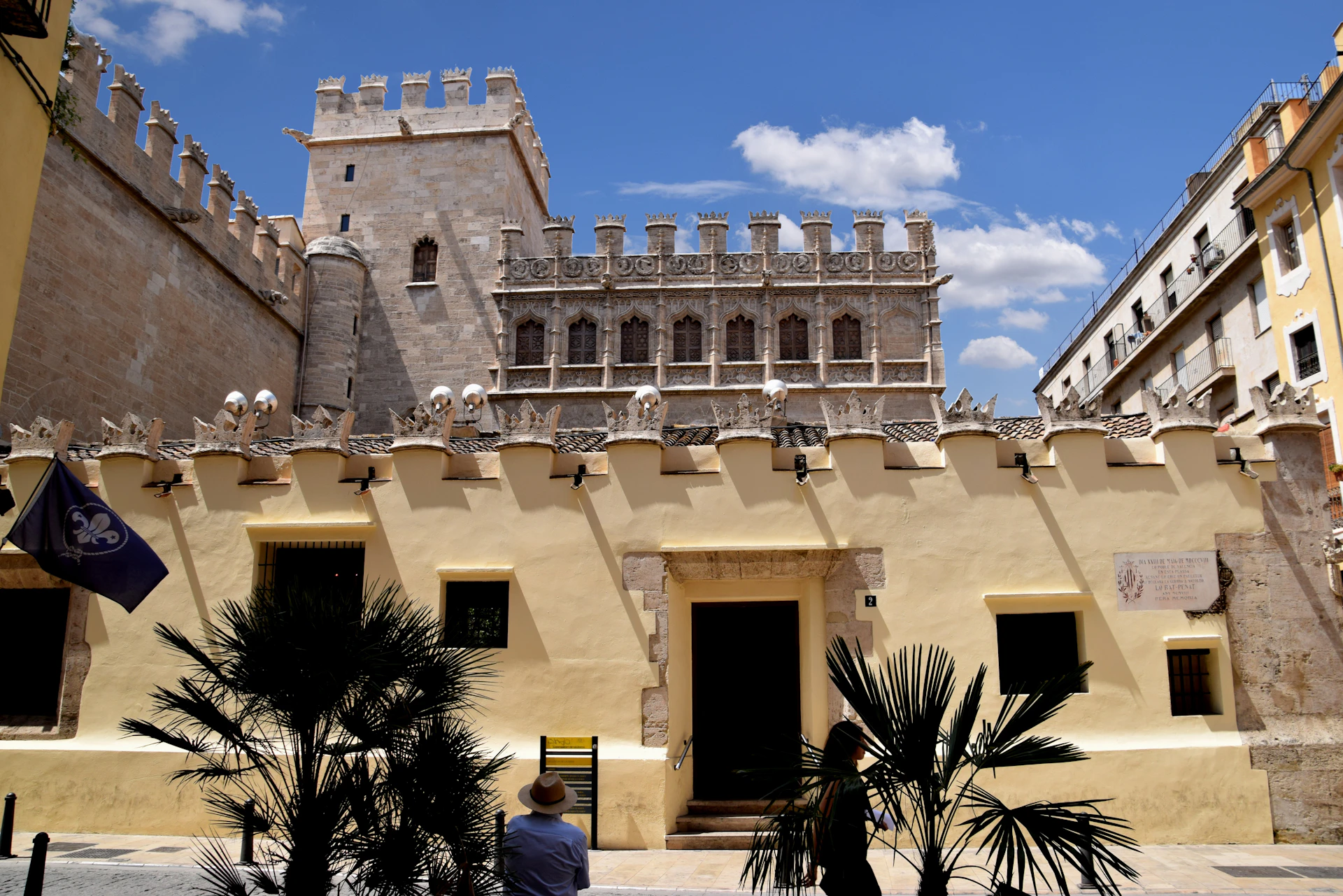 Historic fortress with crenelated walls and a central doorway, silhouetted figures, and palm trees in the foreground under a blue sky.