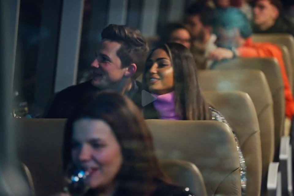 People sitting on a bus in soft lighting, smiling and conversing. The focus is on a man and a woman in the middle of the image.