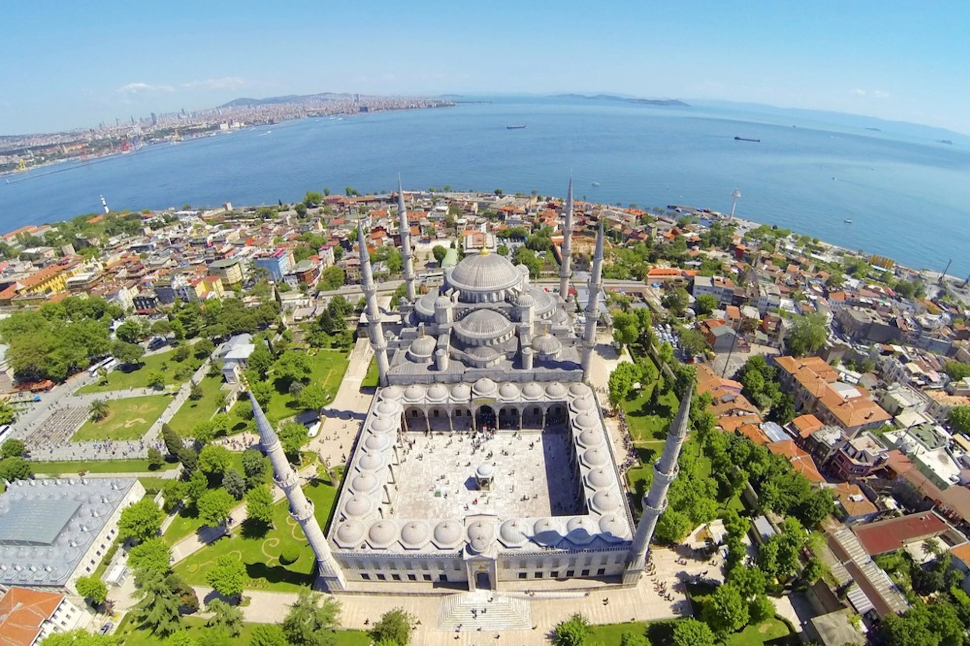 Aerial view of a large mosque with multiple domes and minarets, surrounded by trees and buildings, near a body of water.